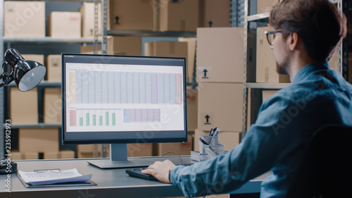 Warehouse Inventory Manager Works with a Spreadsheet on a Personal Computer while Sitting at His Desk. In the Background Shelves Full of Cardboard Box Packages Ready For Shipping. photo