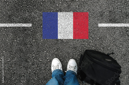  a man with a shoes and backpack is standing on asphalt next to flag of France and border