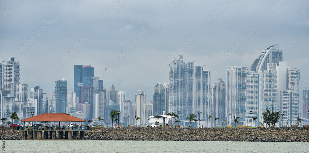 Obraz premium Hot, humid day in Panama city as another rainstorm brews quickly over the city skyline. Tall buildings shimmer in heatwaves rising in humid air. People on Panama Canal jetty park in foreground.