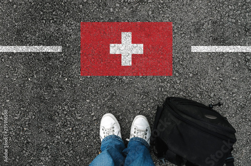 Photography a man with a shoes and backpack is standing on asphalt nex to flag of Switzerla