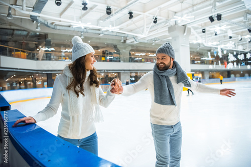 smiling couple in hats and sweaters holding hands while skating on ice rink