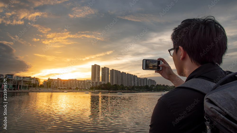 Asian man with glasses using smartphone camera taking photo during the sunset in the city. Urban lifestyle with mobile phone technology concept