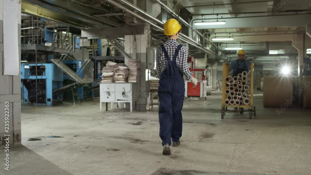 Female technician in overall and hardhat taking cart with paper reels from male coworker and pulling it while mid-aged supervisor writing on clipboard in printing plant