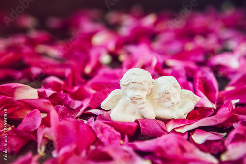 Statuette of two antique little lovely angels of the gypsum on the black stone background with pink purple peony flower petals. Love, romantic, Valentines day concept. Close up, soft selective focus.