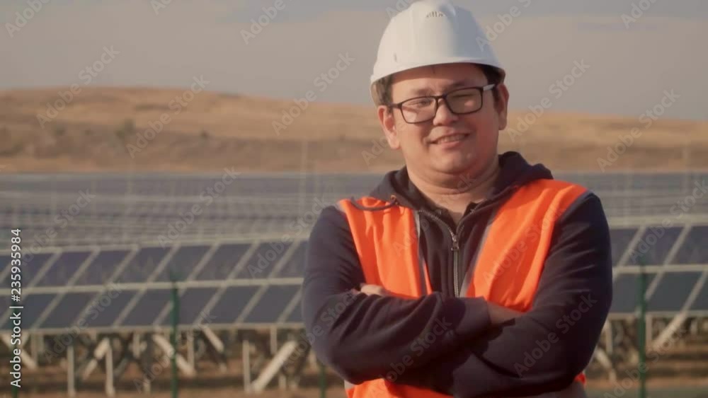 Asian man in an engineer suit on the background of a solar power plant looking at the horizon.