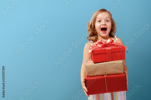 Happy little blonde child girl holding three boxes with gifts on a blue isolated background. The concept of celebrating, giving and receiving a gift