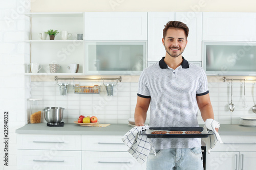 Young man holding oven sheet with cookies in kitchen, space for text