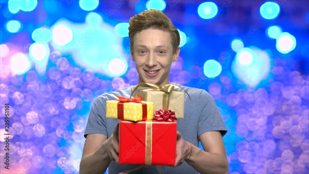 Teen guy giving Christmas gifts. Portrait of cute teen boy handing Christmas gifts to camera on blue bokeh background.