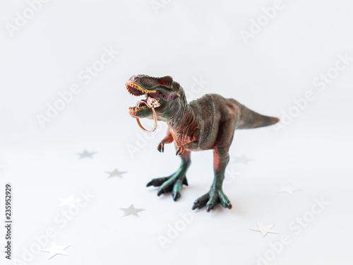 Dinosaur with engagement ring. Plastic toy with wedding jewelry. White background with star confetti. © Konstantin Aksenov