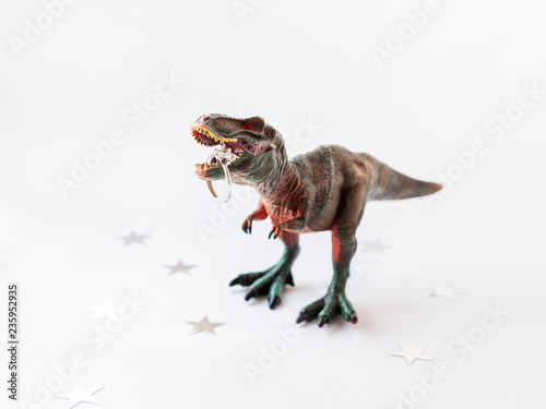 Dinosaur with engagement ring. Plastic toy with wedding jewelry. White background with star confetti.