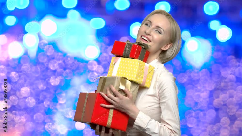 Woman holding stack of Christmas gift boxes. Cheerful girl with pile of gift boxes on blue bokeh background. Winter holidays concept.