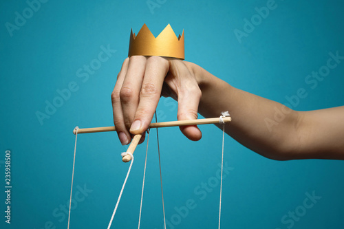 Concept of manipulation. Hand with crown holds strings for manipulation on blue background.