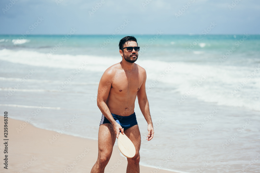 Young Brazilian man playing beach tennis on the beach with swimwear Young Brazilian man playing beach tennis on the beach with swimwear