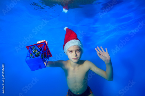 Portrait of a little boy who swims and poses under the water in a red hat of Santa Claus with a toy in his hand. Landscape orientation