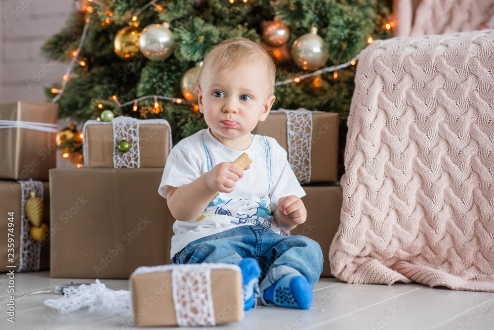 Little blond boy eating a gingerbread man by the Christmas tree at home. New Year holiday card with garlands bokeh. The baby laughs, opens a gift box from Santa Claus. Waiting for miracle. Copy space