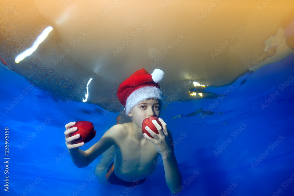 A child in a red Christmas cap floats under water with red apples in his hands on a blue background. He bites an apple and looks at the camera. Portrait. Close up. Horizontal view A child in a red Christmas cap floats under water with red apples in his hands on a blue background. He bites an apple and looks at the camera. Portrait. Close up. Horizontal view