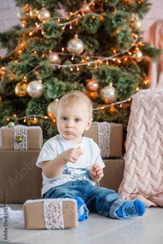 Little blond boy eating a gingerbread man by the Christmas tree at home. New Year holiday card with garlands bokeh. The baby laughs  opens a gift box from Santa Claus. Waiting for miracle. Copy space