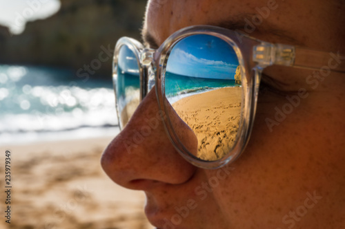Young woman or girl watches a sea on the beach and scene is mirroring in her sunglasses, Benagil beach, Portugal, popular travel destination in Europe