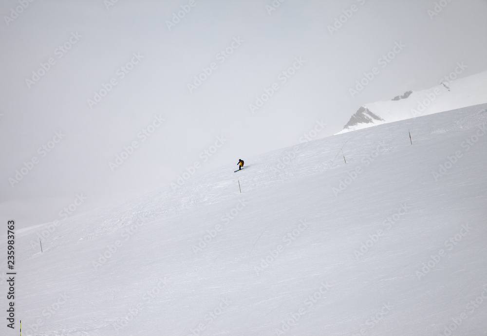 Skier downhill on snowy off-piste ski slope and mountains in fog