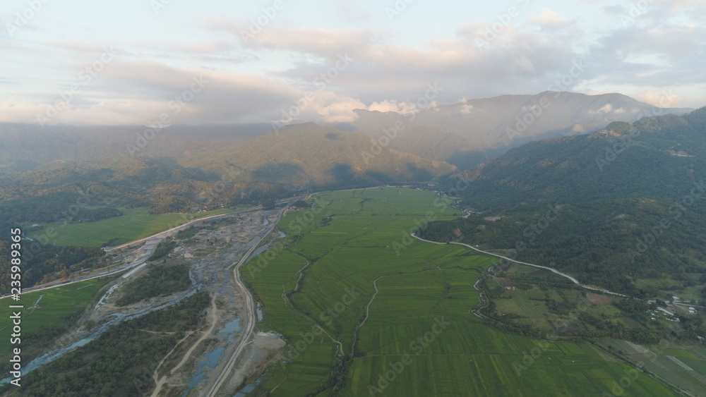 Fototapeta premium Aerial view of farmer fields, river, village in the background of mountains at sunset on the island of Luzon, Philippines. Mountain landscape with valley and village.