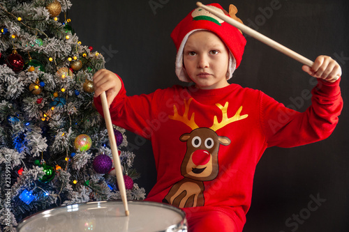 Happy Caucasian childish drummer in carnival costume Santa Claus Deer plays on a new drum set with drum sticks in his hands. Parents bought child drum as gift for Christmas. Lights on Christmas tree