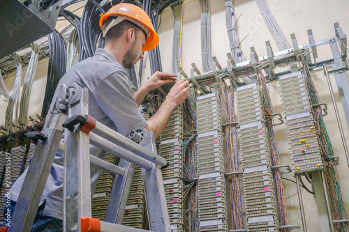  The engineer works in the server room of the data center. Cable laying in telephone exchange. A worker standing on the stairs commutes telephone wires.