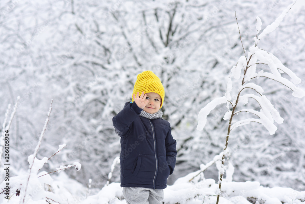 Hello, winter! Handsome boy in black warm jacket and yellow knitted cap. Kid in snow waving hand hello. Friendly boy smiles in the winter forest. Happy childhood. Positive children. Winter walk