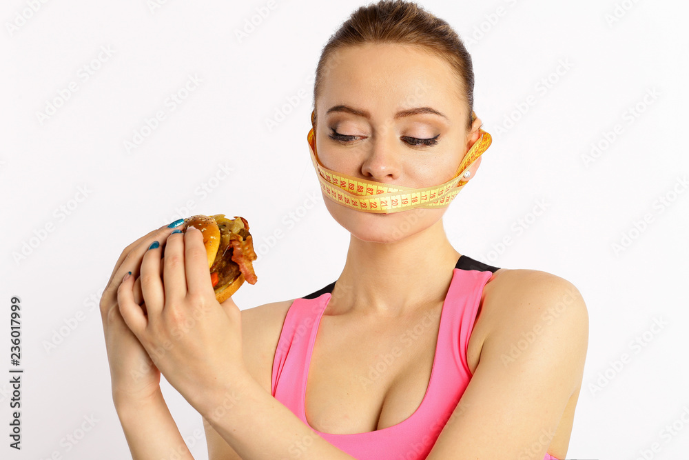 Young and beautiful woman and measuring tape holding a high-calorie hamburger isolated white background. The concept of avoiding harmful fast foods and diet Young and beautiful woman and measuring tape holding a high-calorie hamburger isolated white background. The concept of avoiding harmful fast foods and diet
