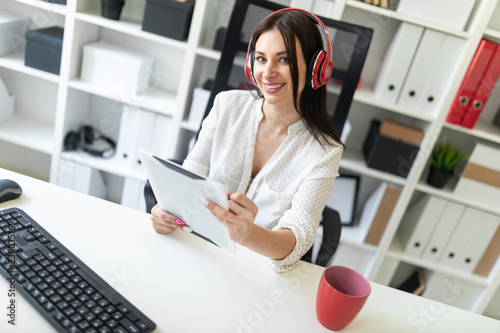 A young girl with headphones sitting in the office at the computer table and holding a tablet.