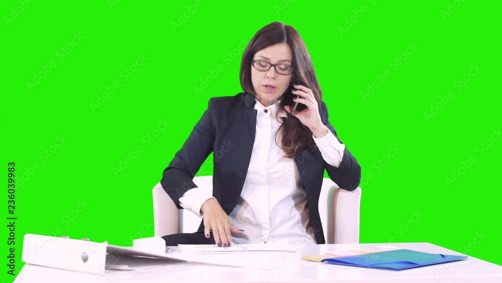 Young brunette on a green background sits behind a Desk in office and talking on the phone. On the table are documents and the contract. Young brunette on a green background sits behind a Desk in office and talking on the phone. On the table are documents and the contract.