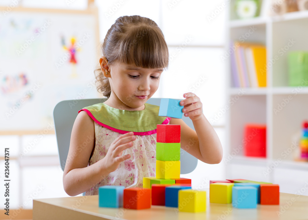 Little child girl plays with wooden colorful cubes sitting at table in nursery room or kindergarten Little child girl plays with wooden colorful cubes sitting at table in nursery room or kindergarten