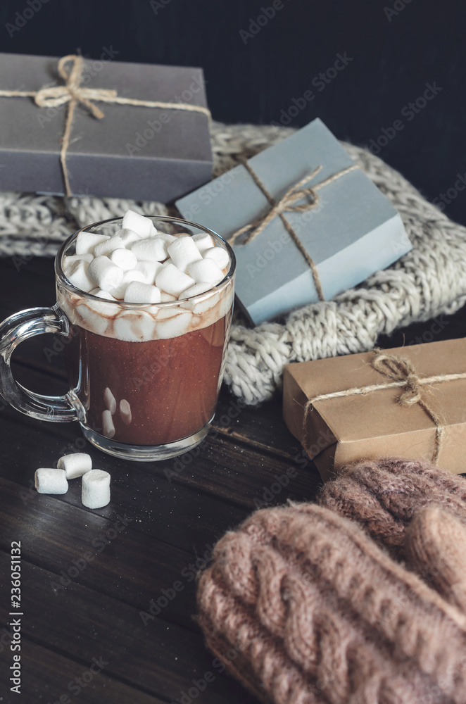 Hot cocoa with marshmallows in a glass cup surrounded by Christmas gifts and winter clothes, Christmas atmosphere and coziness