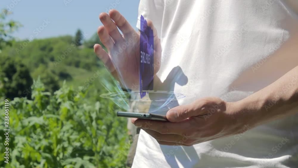 Man shows concept hologram Explore on his phone. Person in white t-shirt with future technology holographic screen and green nature background Man shows concept hologram Explore on his phone. Person in white t-shirt with future technology holographic screen and green nature background
