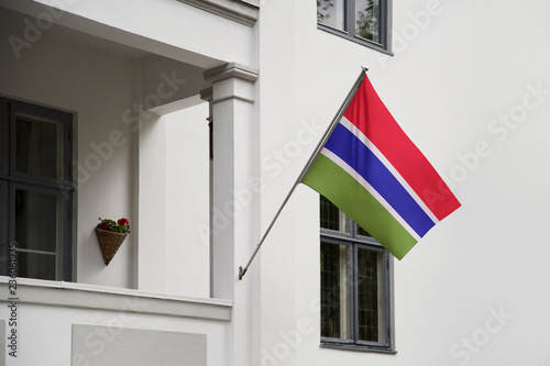 Gambia flag hanging on a pole in front of the house. National flag waving on a home displaying on a pole on a front door of a building and raised at a full staff.
