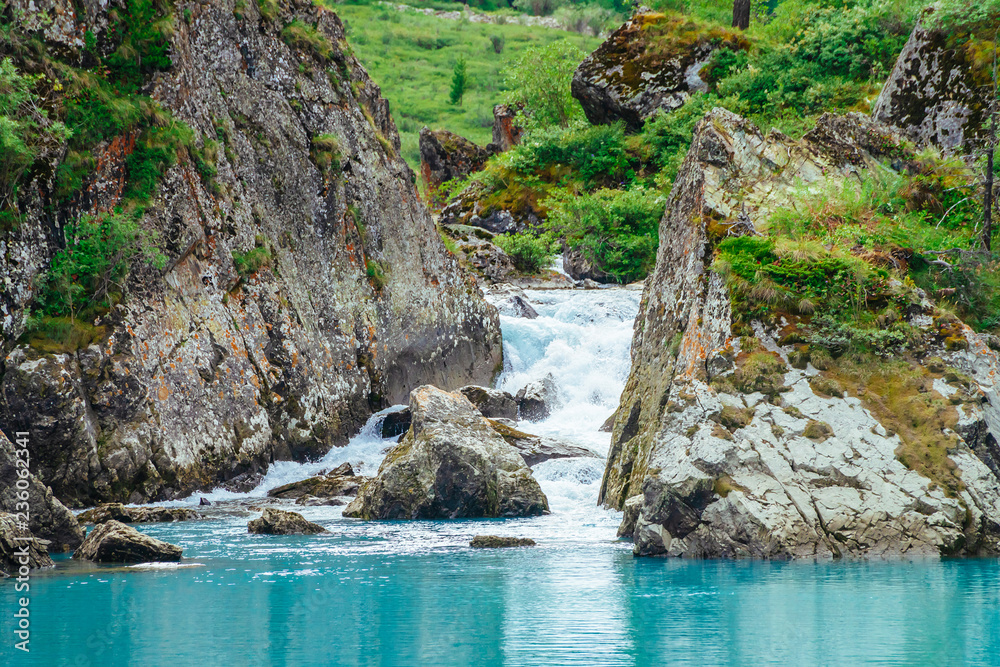 Mountain creek flows into azure lake. Fast stream of brook. Smooth water surface. Wonderful rocks with rich vegetation of highlands. Amazing landscape of majestic nature.