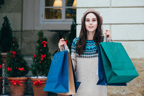 Outdoor portrait of smiling woman posing on street, holding paper shopping bags, looking at camera, wearing stylish clothes. Winter holidays, Christmas, New Year concept.