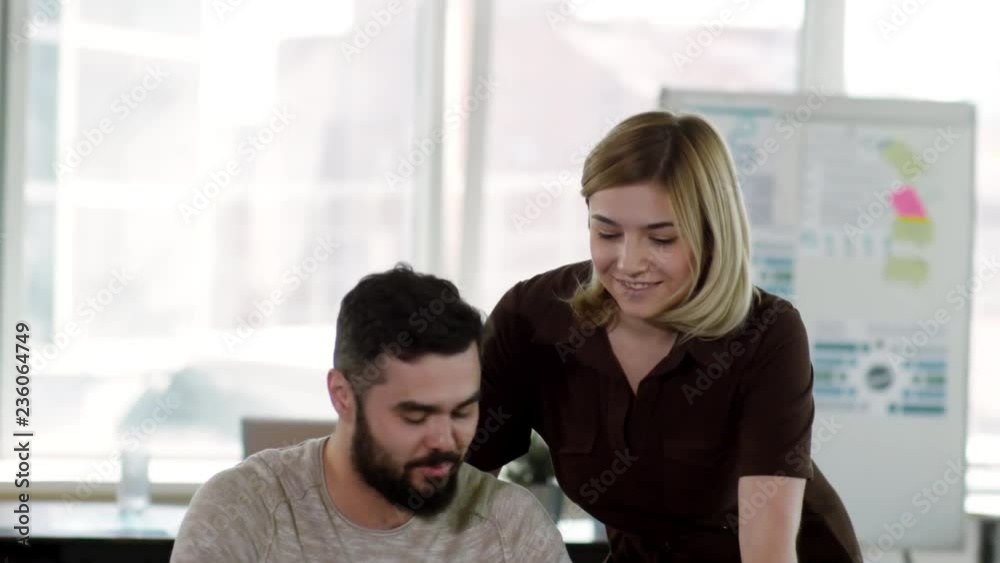 Tilt down of young Caucasian woman standing by male colleague at desk, pointing at laptop screen and explaining him something while working together in office Tilt down of young Caucasian woman standing by male colleague at desk, pointing at laptop screen and explaining him something while working together in office