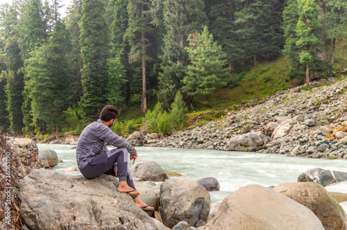 A person sitting on a rock in front of a stream with forest in background at Pahalgam in Kashmir