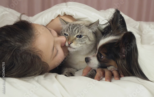 Happy teen girl communicates with dog Papillon and Thai cat in bed