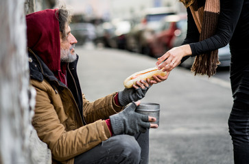 Unrecognizable woman giving food to homeless beggar man sitting in city.