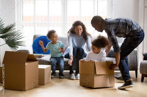 Happy playful large African American family moving in new apartment, little preschooler daughter sitting in cardboard boxes, father rolling her to mother, playing together, purchase property concept