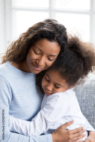 Loving African American mother embracing with preschooler little adorable daughter, sitting together on couch at home, warm relationships parent and child, closeness, love and support concept vertical