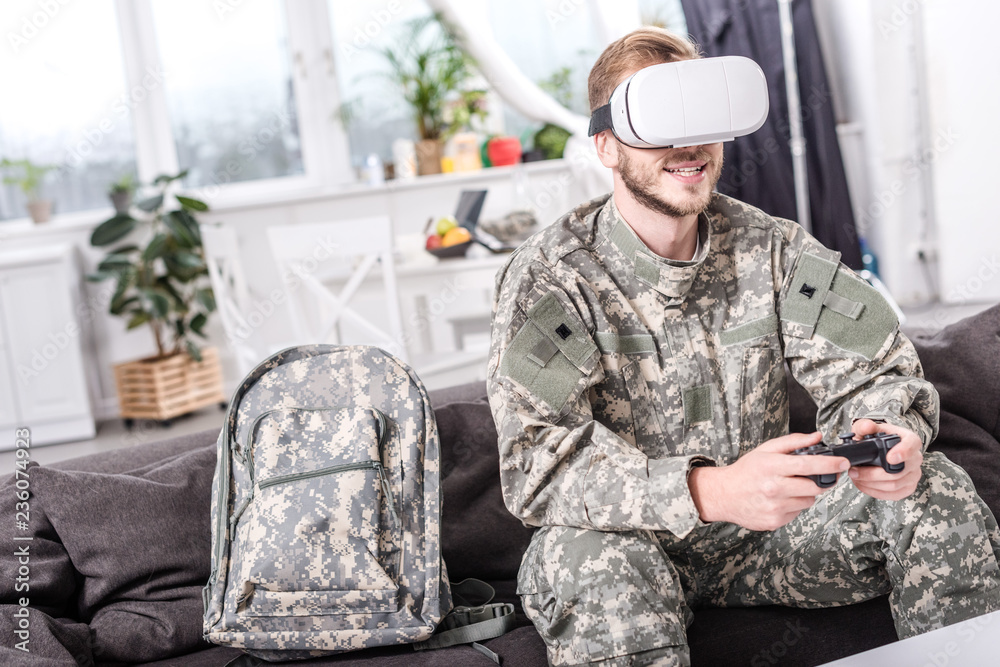 Fototapeta premium smiling soldier in virtual reality headset playing video game on couch