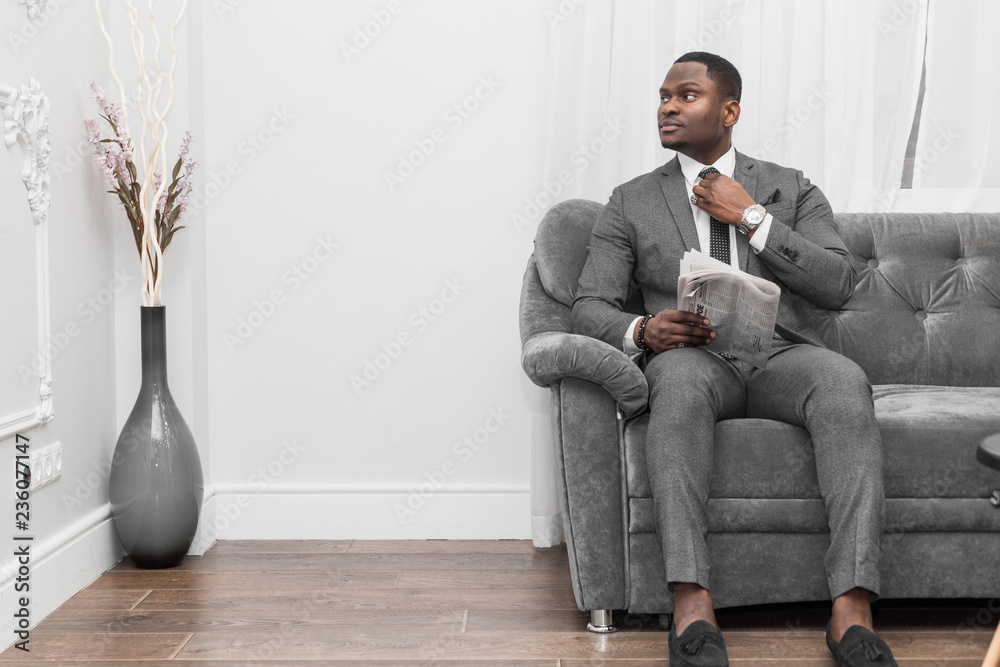 Young African American businessman in a gray suit reading a newspaper while sitting on a sofa. Young African American businessman in a gray suit reading a newspaper while sitting on a sofa.
