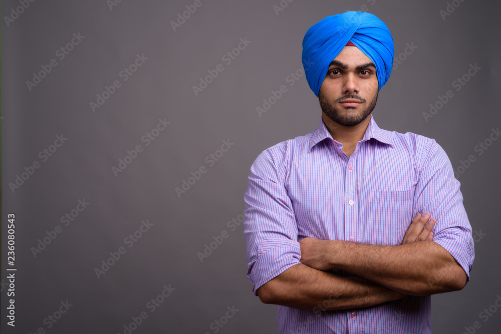 Young handsome Indian businessman wearing turban against gray ba