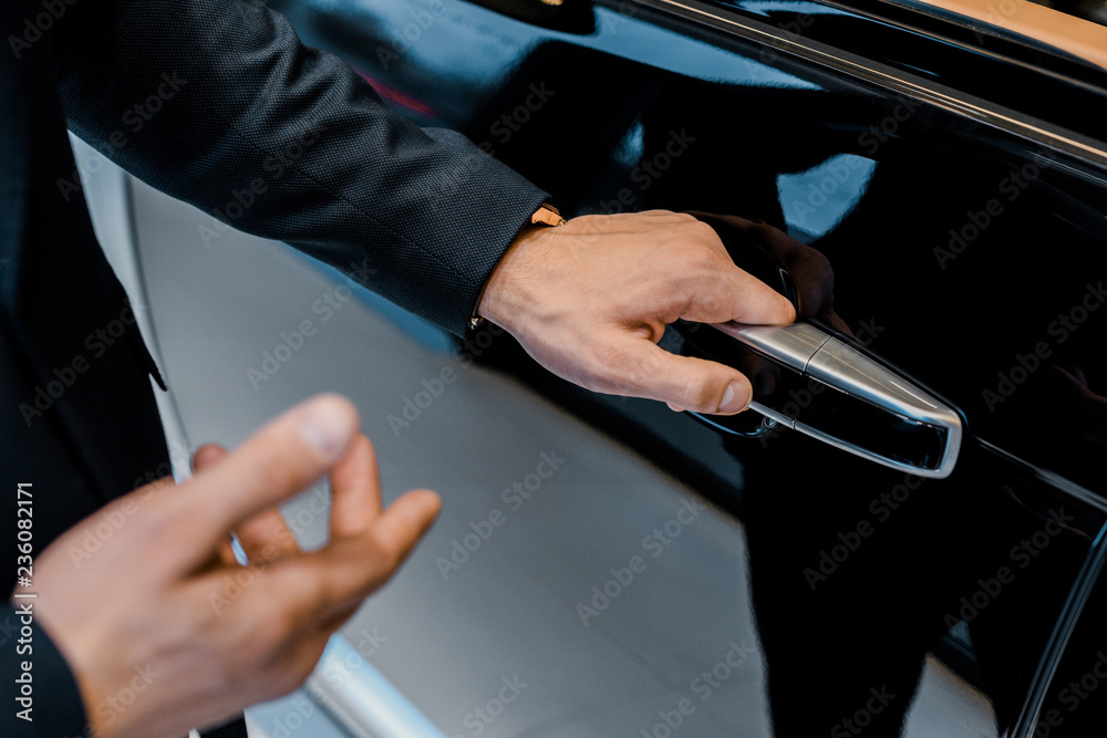 partial view of male car dealer opening door and pointing at black automobile in car salon