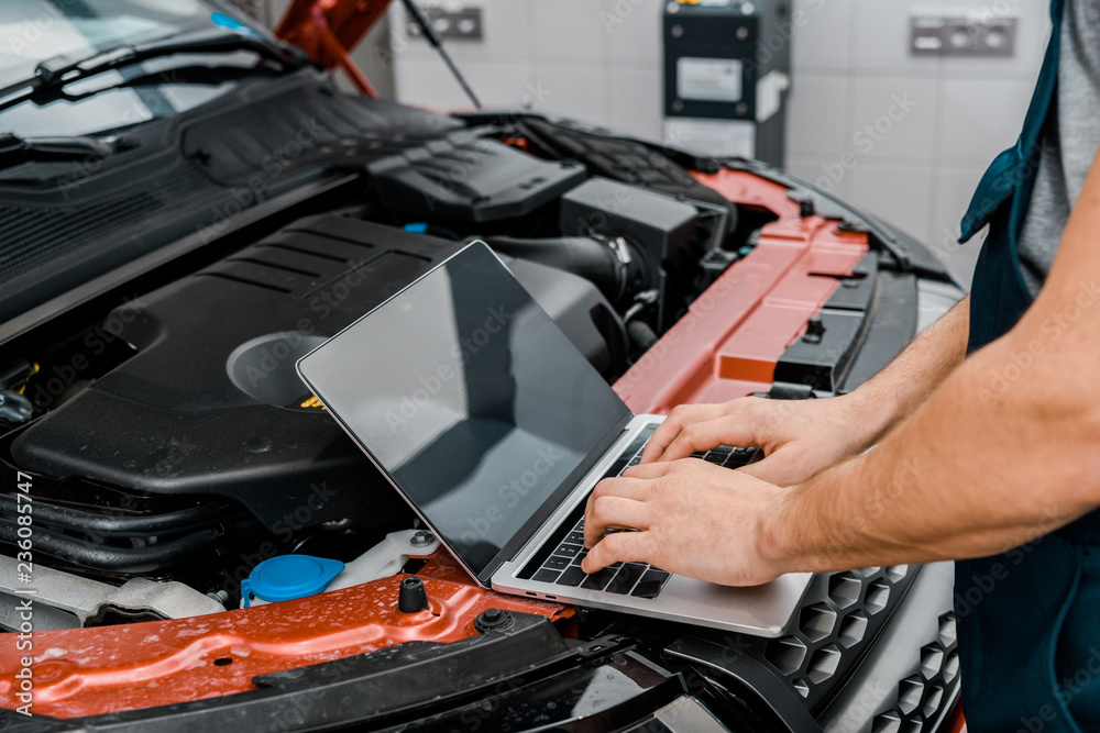 cropped shot of auto mechanic working on laptop with blank screen at automobile with opened car cowl at mechanic shop