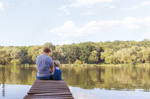 Dad and a tiny girl in dungarees relax at the river. Back view, copy space.