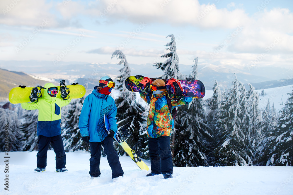 Three snowboarders walking at ski resort. Friends climbing to mountain top carrying their snowboards through forest for backcountry freeride and wearing reflective goggles, colorful fashion clothes.