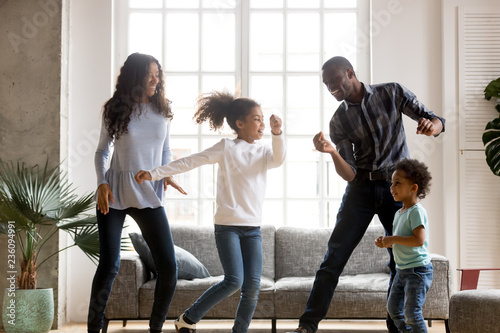 Happy African American having fun together indoors, funny married couple dancing with adorable little preschooler daughter and cute toddler son at home, listening to music, family weekend with kids